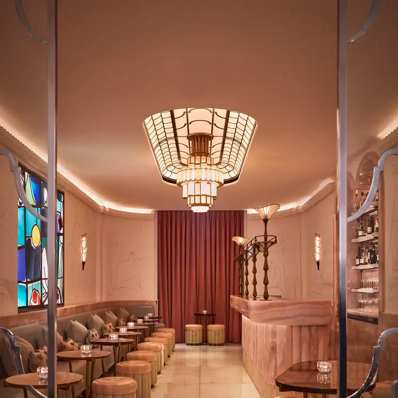 Art Deco–inspired bar with blush pink marble counter, upholstered stools, and soft lighting from a geometric chandelier. A stained-glass window and deep red curtains add warmth and elegance to the intimate space.