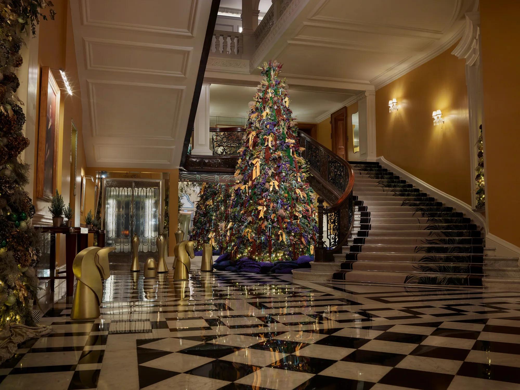 A colourful Christmas tree stands beneath a sweeping staircase in a grand hotel lobby with patterned marble floors and festive greenery.