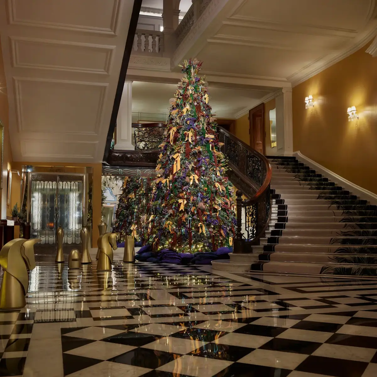 A colourful Christmas tree stands beneath a sweeping staircase in a grand hotel lobby with patterned marble floors and festive greenery.