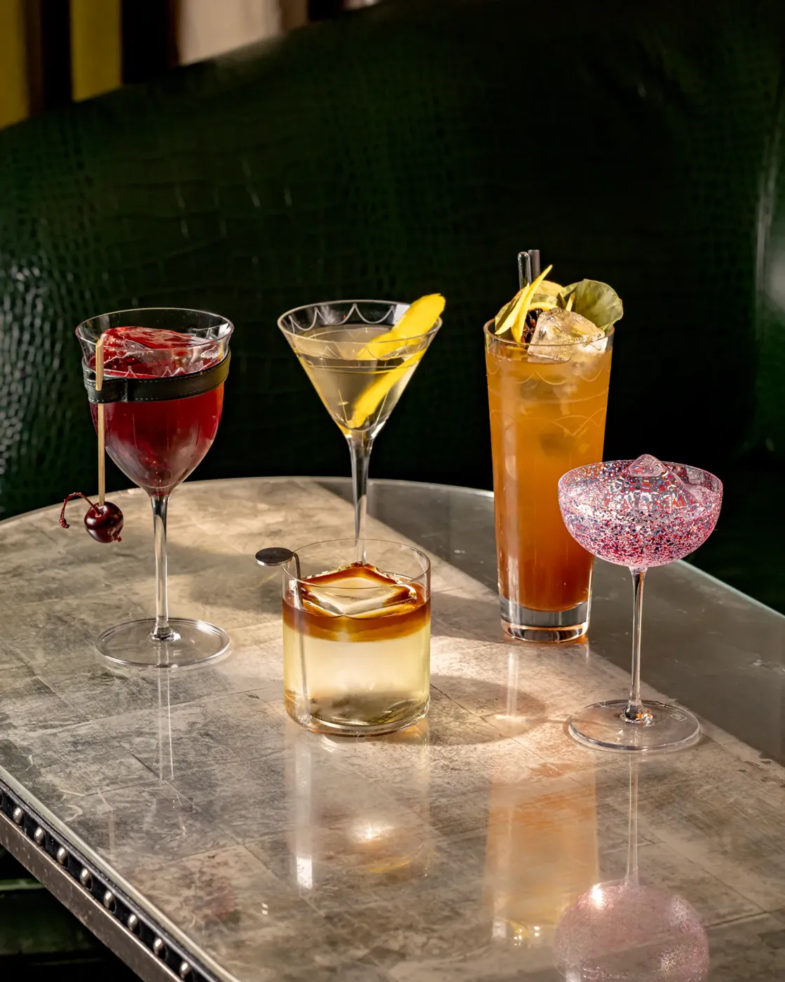Five colorful cocktails arranged in a row on a bar, each garnished with fruit and herbs.