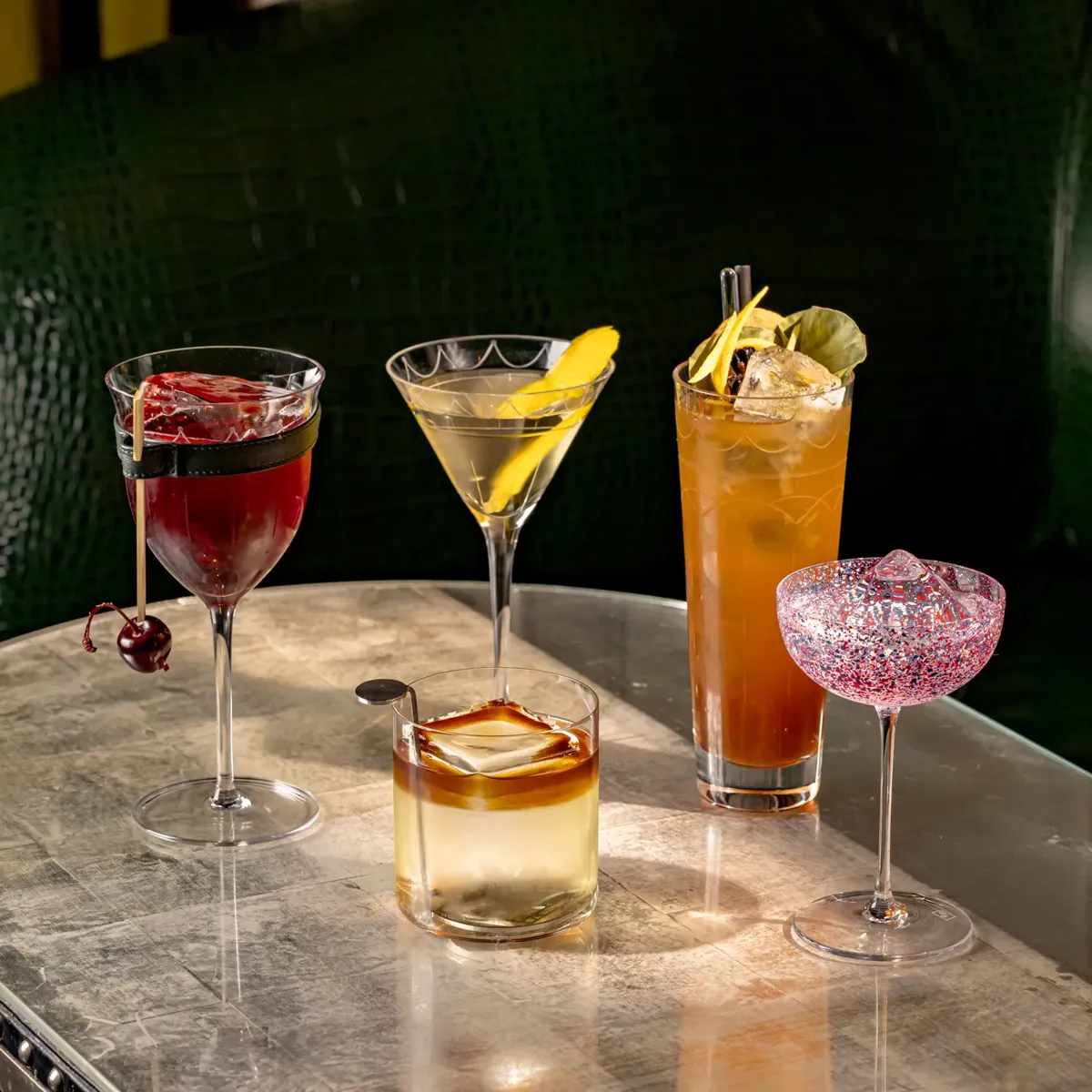 Five colorful cocktails arranged in a row on a bar, each garnished with fruit and herbs.