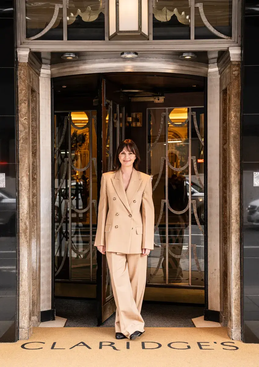 A woman in a beige suit stands smiling at the entrance of Claridge’s, framed by the hotel’s revolving doors and Art Deco detailing.