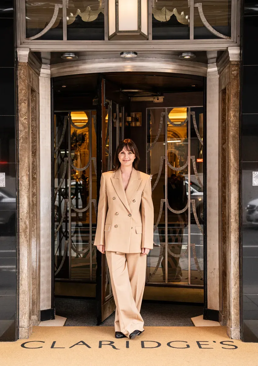 A woman in a beige suit stands smiling at the entrance of Claridge’s, framed by the hotel’s revolving doors and Art Deco detailing.