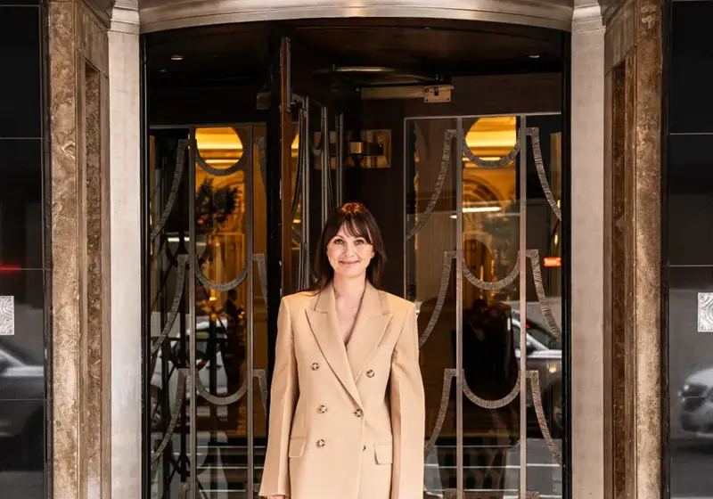 A woman in a beige suit stands smiling at the entrance of Claridge’s, framed by the hotel’s revolving doors and Art Deco detailing.