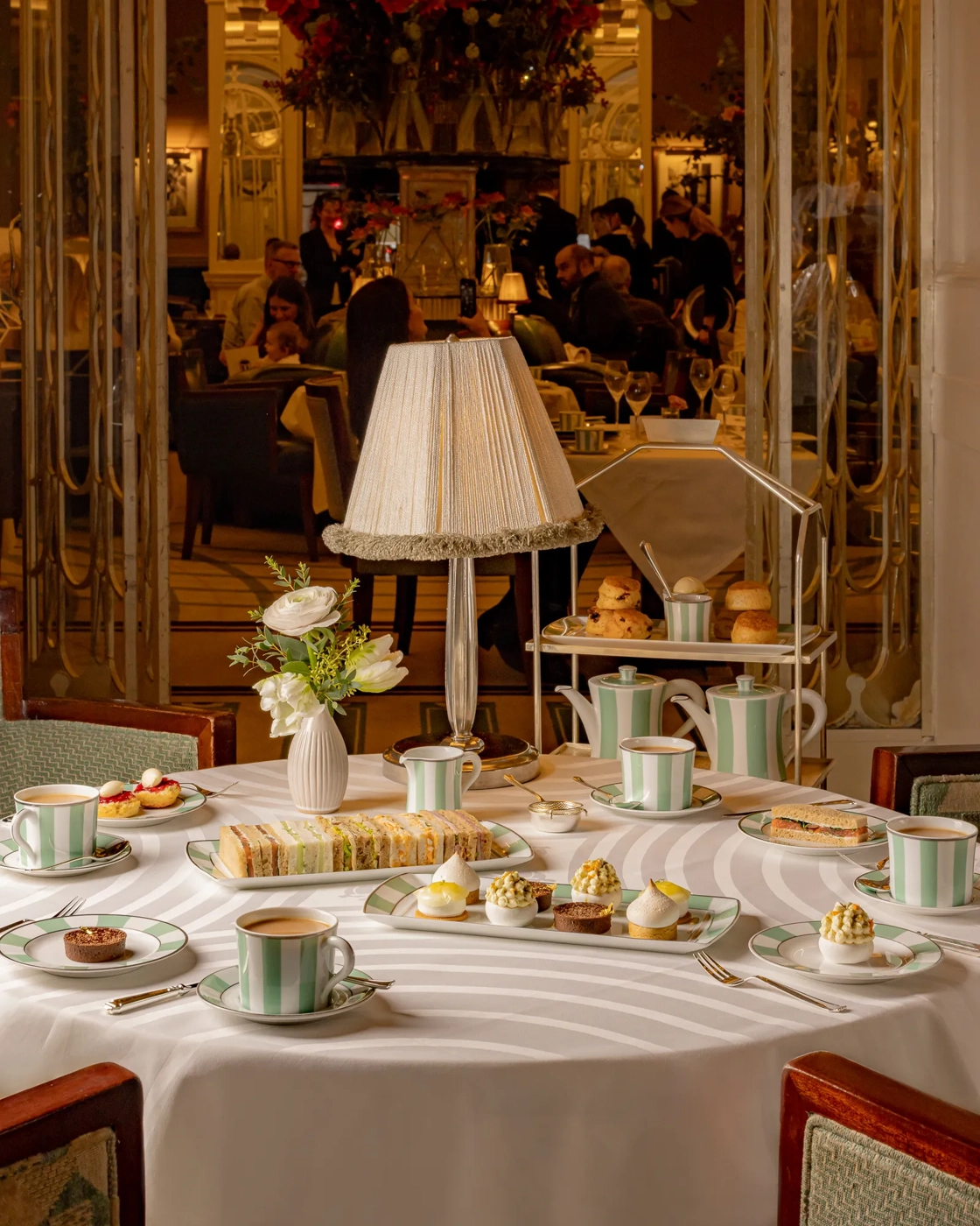 Afternoon tea table at Claridge’s set with striped china, pastries, sandwiches, and a tiered stand beneath a soft-lit table lamp.