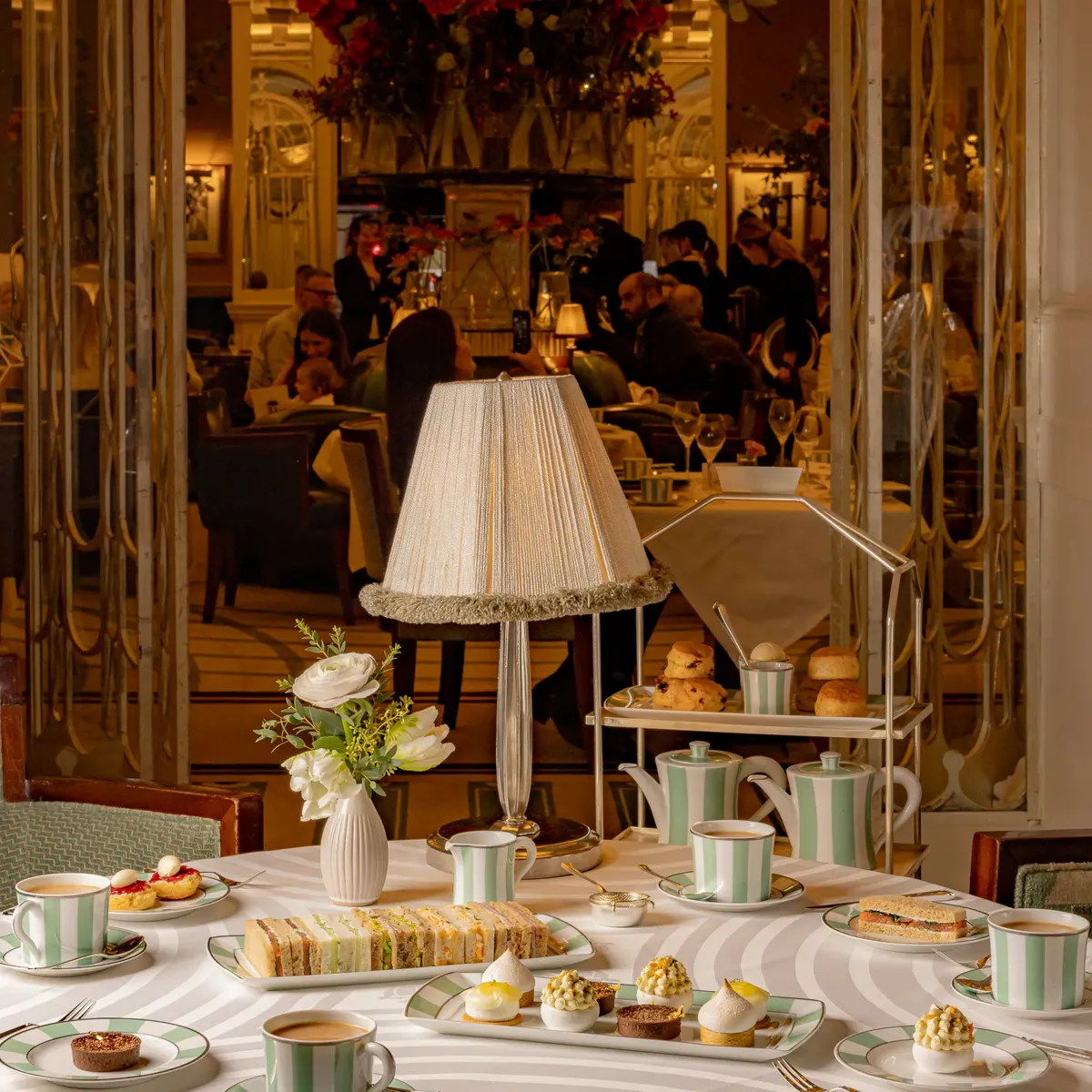 Afternoon tea table at Claridge’s set with striped china, pastries, sandwiches, and a tiered stand beneath a soft-lit table lamp.