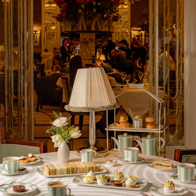 Afternoon tea table at Claridge’s set with striped china, pastries, sandwiches, and a tiered stand beneath a soft-lit table lamp.