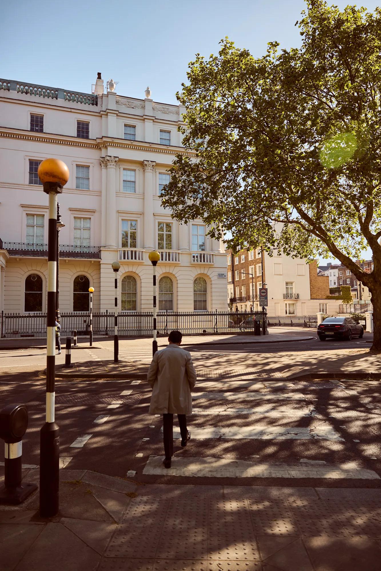 Person crossing a quiet street toward a white neoclassical building, shaded by a large tree on a sunny day.