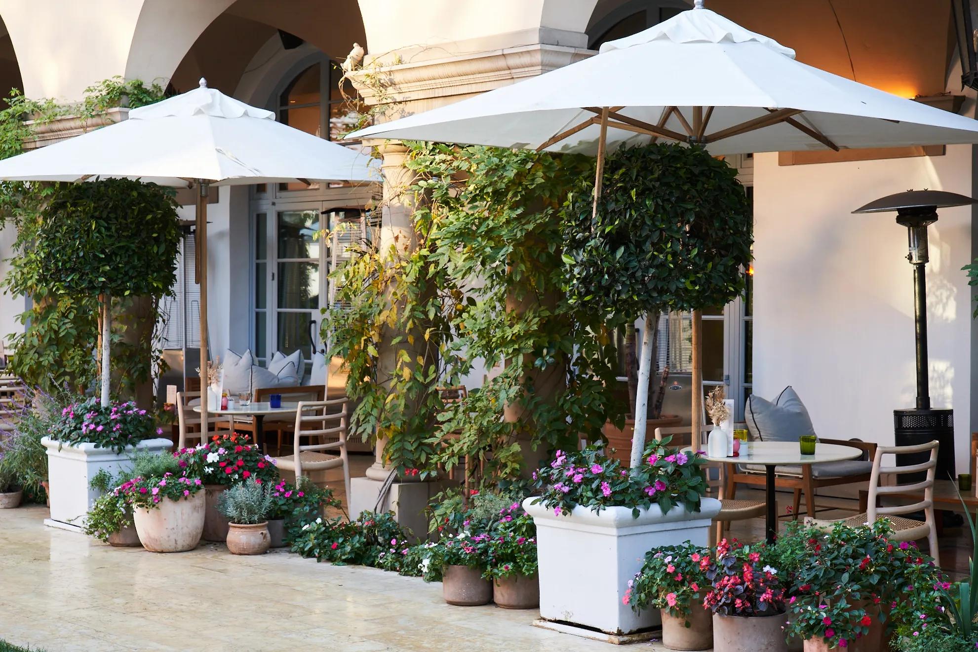 Elegant outdoor terrace with white umbrellas, lush greenery, blooming flowers, and intimate seating beneath arched colonnades.