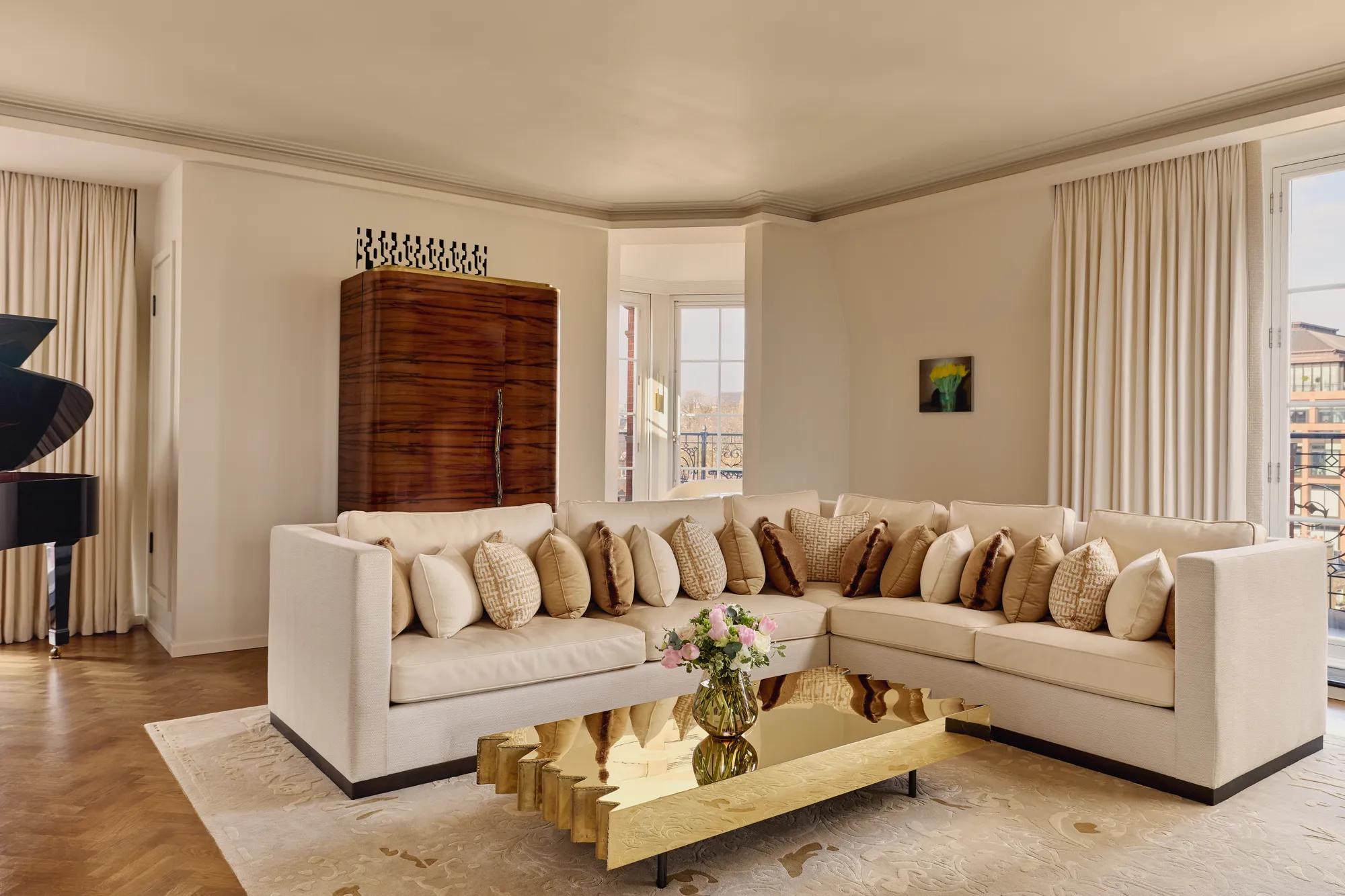 Bright living room with a cream corner sofa adorned with patterned cushions, a sculptural gold coffee table, and large windows framed by light curtains, with a grand piano partially visible in the background.