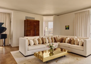 Bright living room with a cream corner sofa adorned with patterned cushions, a sculptural gold coffee table, and large windows framed by light curtains, with a grand piano partially visible in the background.