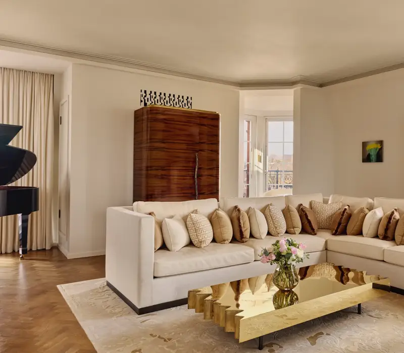 Bright living room with a cream corner sofa adorned with patterned cushions, a sculptural gold coffee table, and large windows framed by light curtains, with a grand piano partially visible in the background.