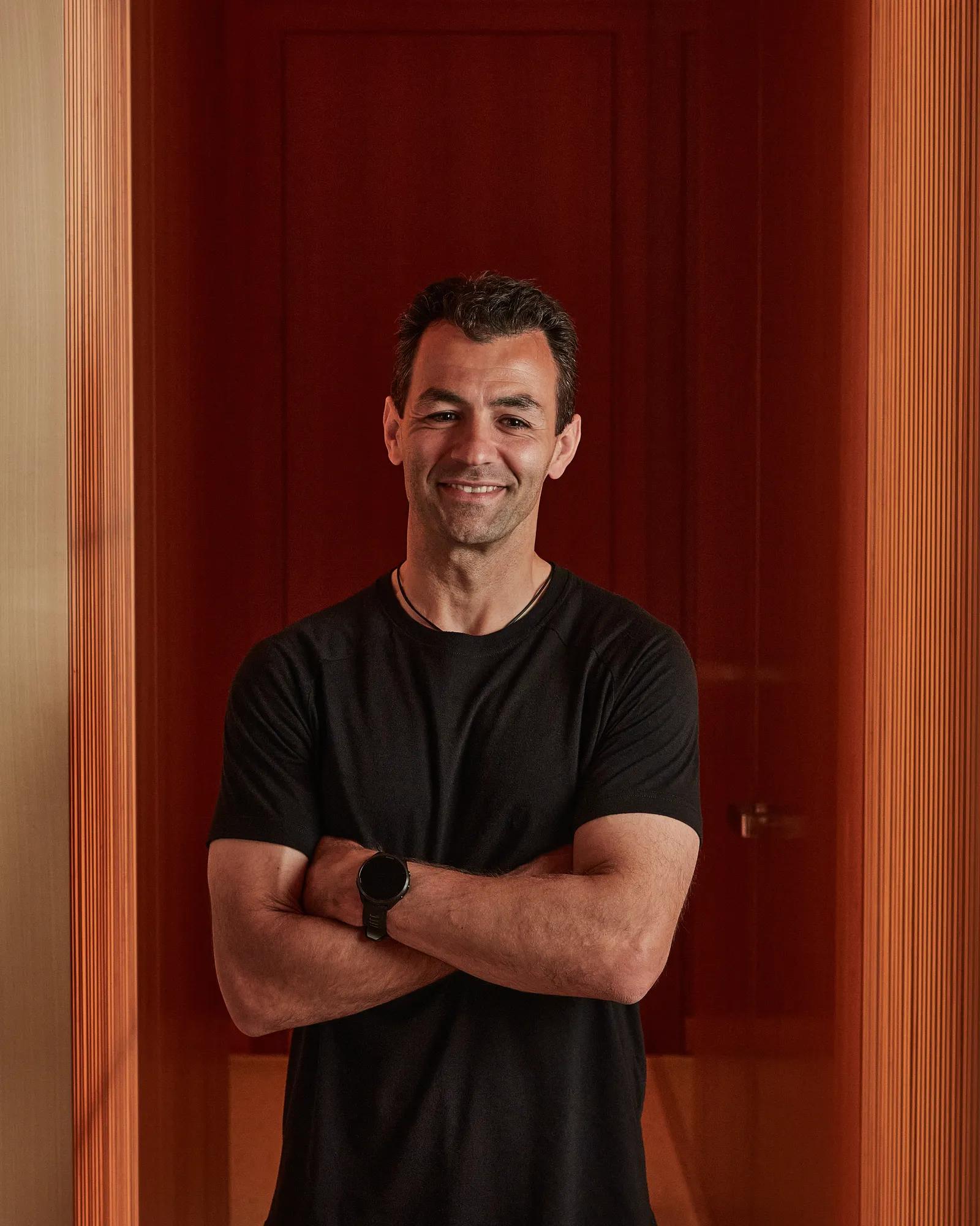 Smiling man in black T-shirt standing with arms crossed against a red wall.