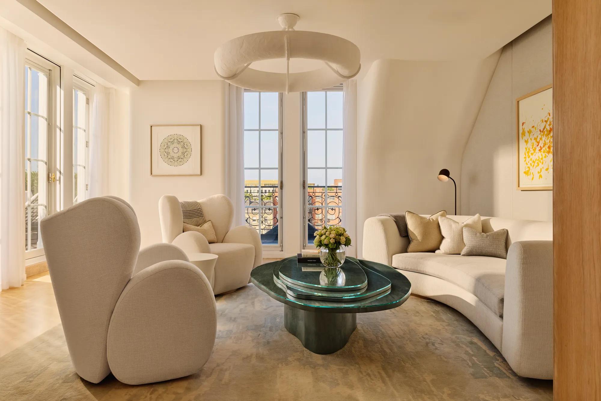 Bright contemporary living room with curved cream armchairs, a matching sofa, and a sculptural green glass coffee table, set beside tall French windows opening onto a terrace with wrought-iron railings.