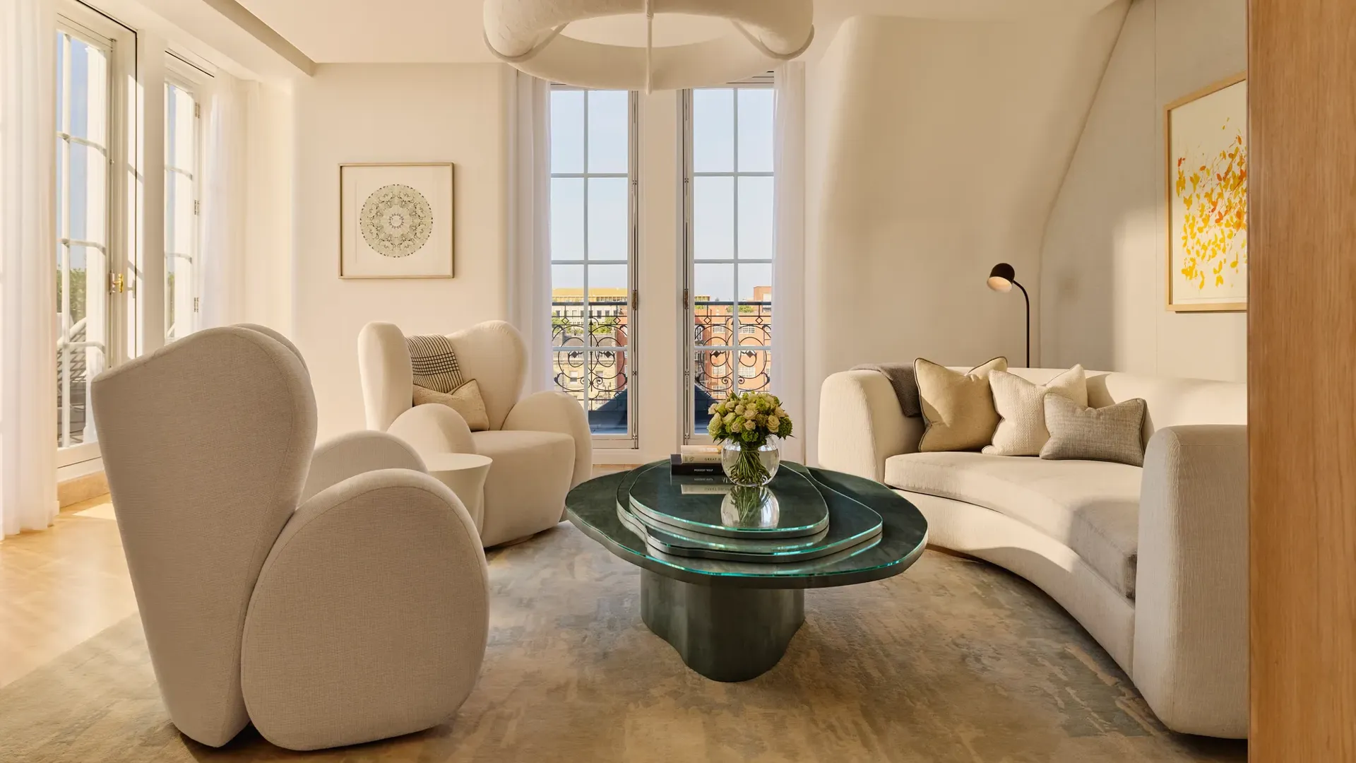 Bright contemporary living room with curved cream armchairs, a matching sofa, and a sculptural green glass coffee table, set beside tall French windows opening onto a terrace with wrought-iron railings.