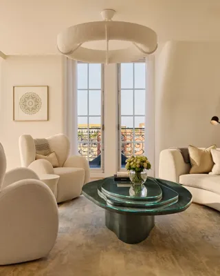 Bright contemporary living room with curved cream armchairs, a matching sofa, and a sculptural green glass coffee table, set beside tall French windows opening onto a terrace with wrought-iron railings.
