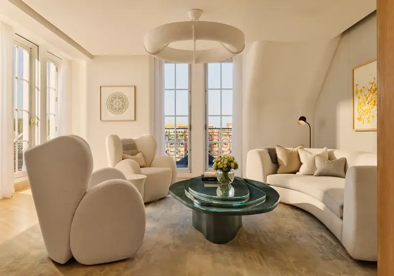 Bright contemporary living room with curved cream armchairs, a matching sofa, and a sculptural green glass coffee table, set beside tall French windows opening onto a terrace with wrought-iron railings.