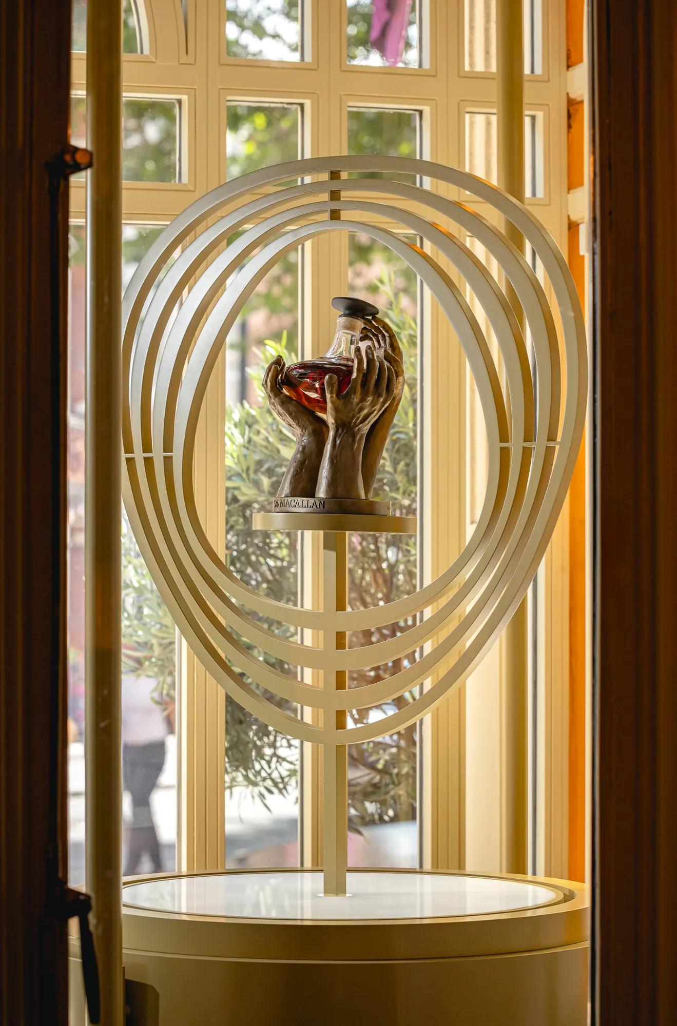 Sculptural display of hands holding a decanter, framed by layered oval rings in front of tall windows.