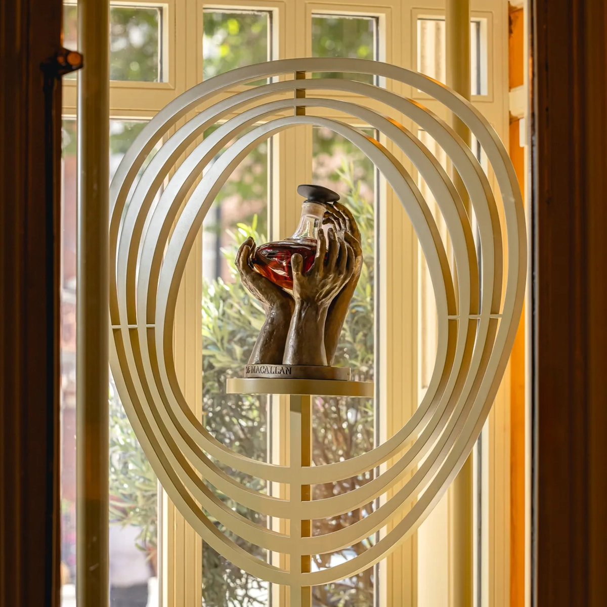 Sculptural display of hands holding a decanter, framed by layered oval rings in front of tall windows.