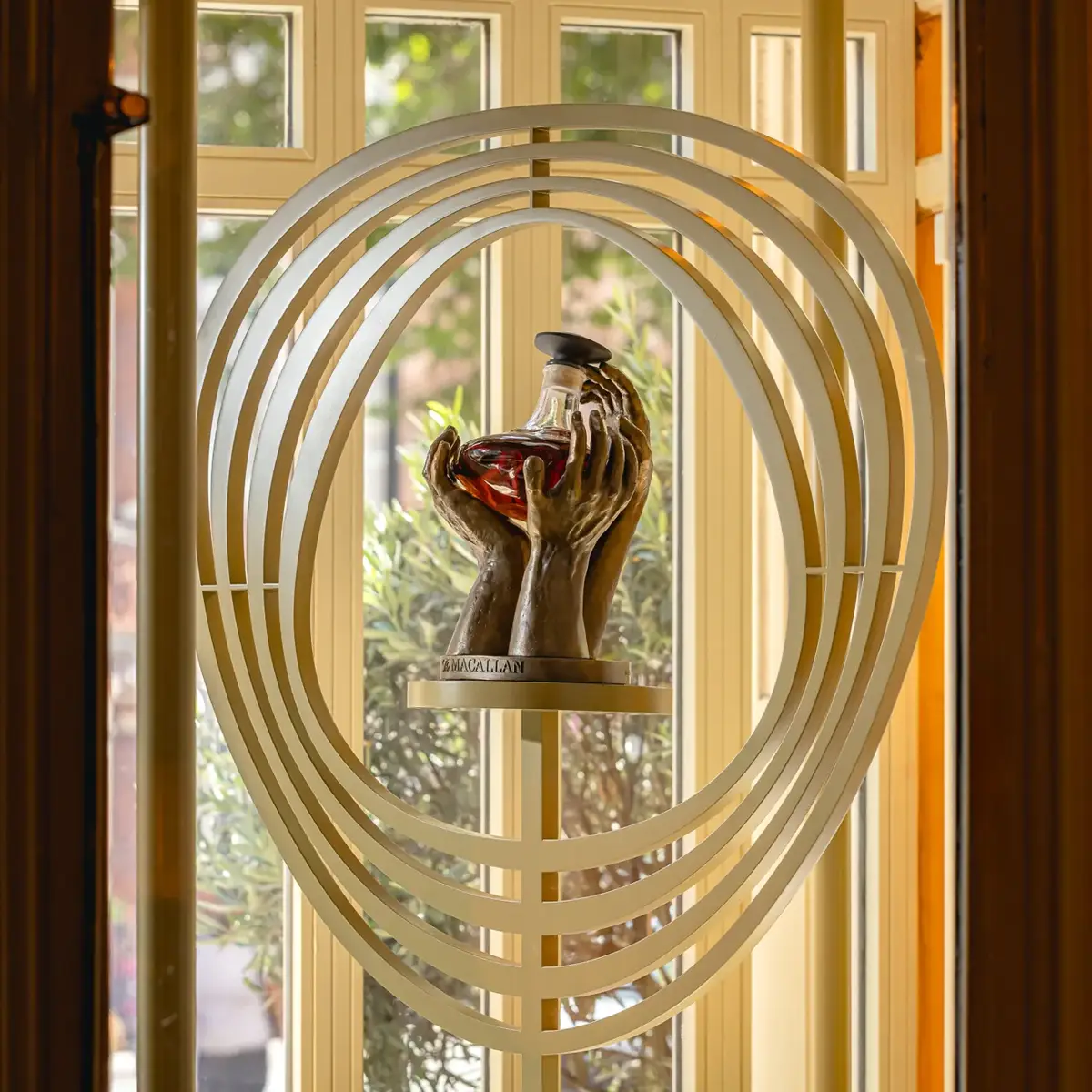 Sculptural display of hands holding a decanter, framed by layered oval rings in front of tall windows.