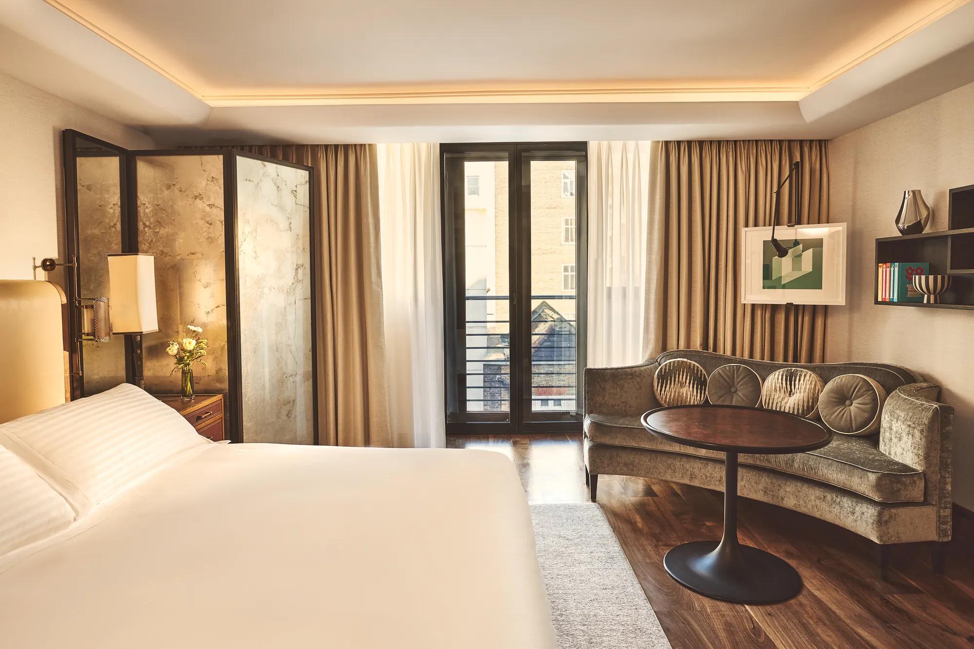 Spacious bedroom with a king-size bed, velvet sofa, walnut floors, curated artwork and French doors opening to a private balcony.
