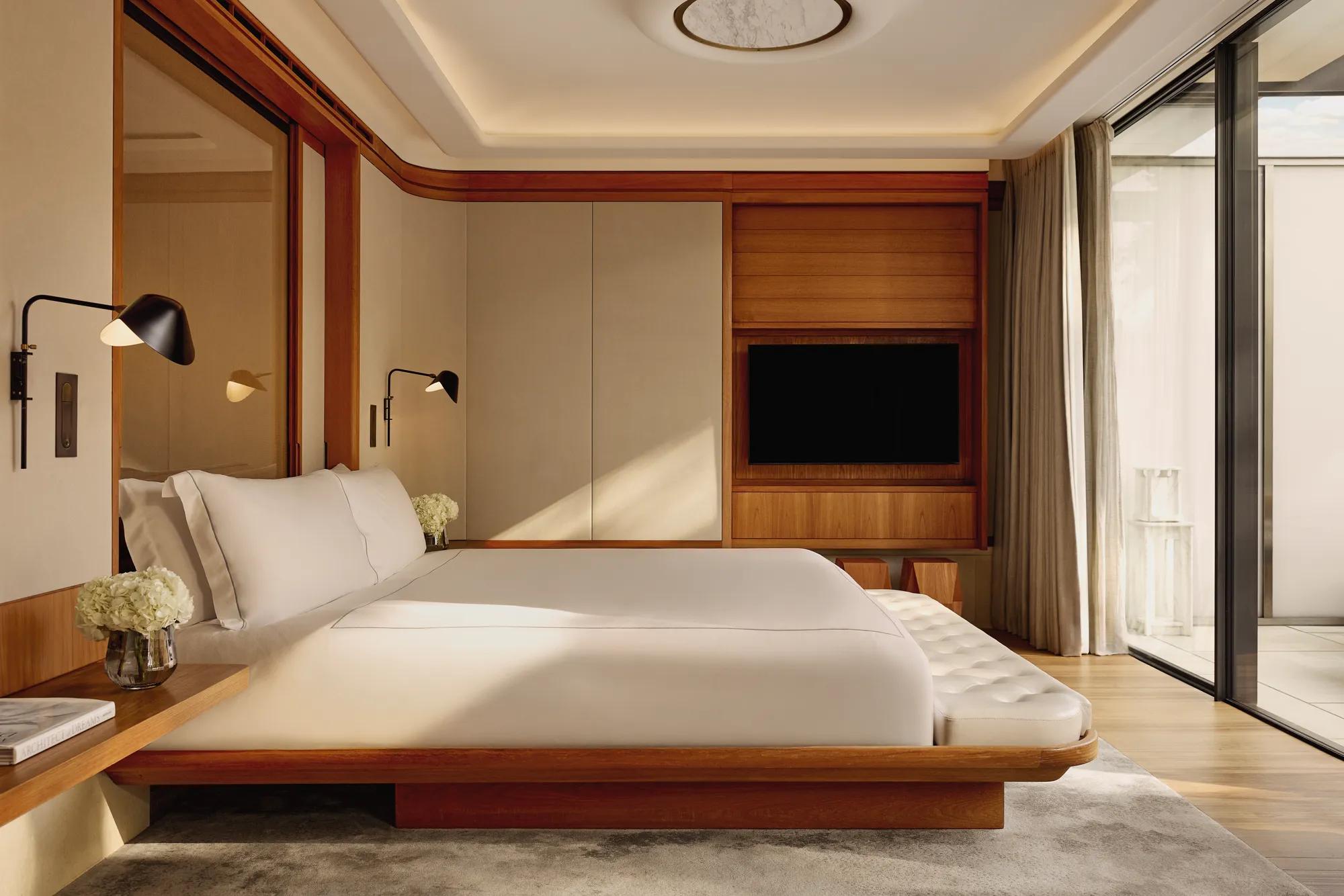 A contemporary bedroom with a low wooden bed frame and crisp white bedding, accented by wall-mounted lamps and large sliding doors opening onto a private terrace.