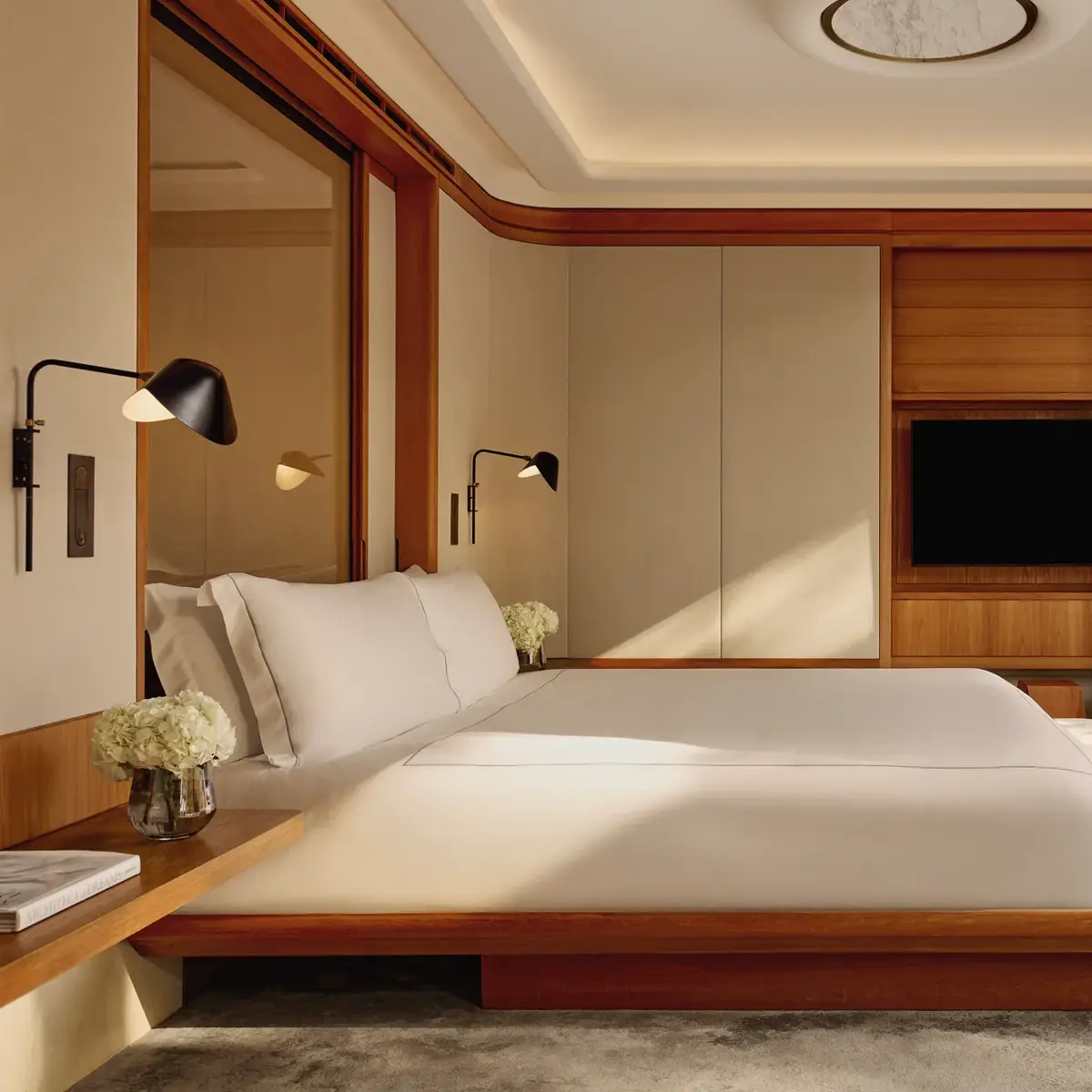 A contemporary bedroom with a low wooden bed frame and crisp white bedding, accented by wall-mounted lamps and large sliding doors opening onto a private terrace.