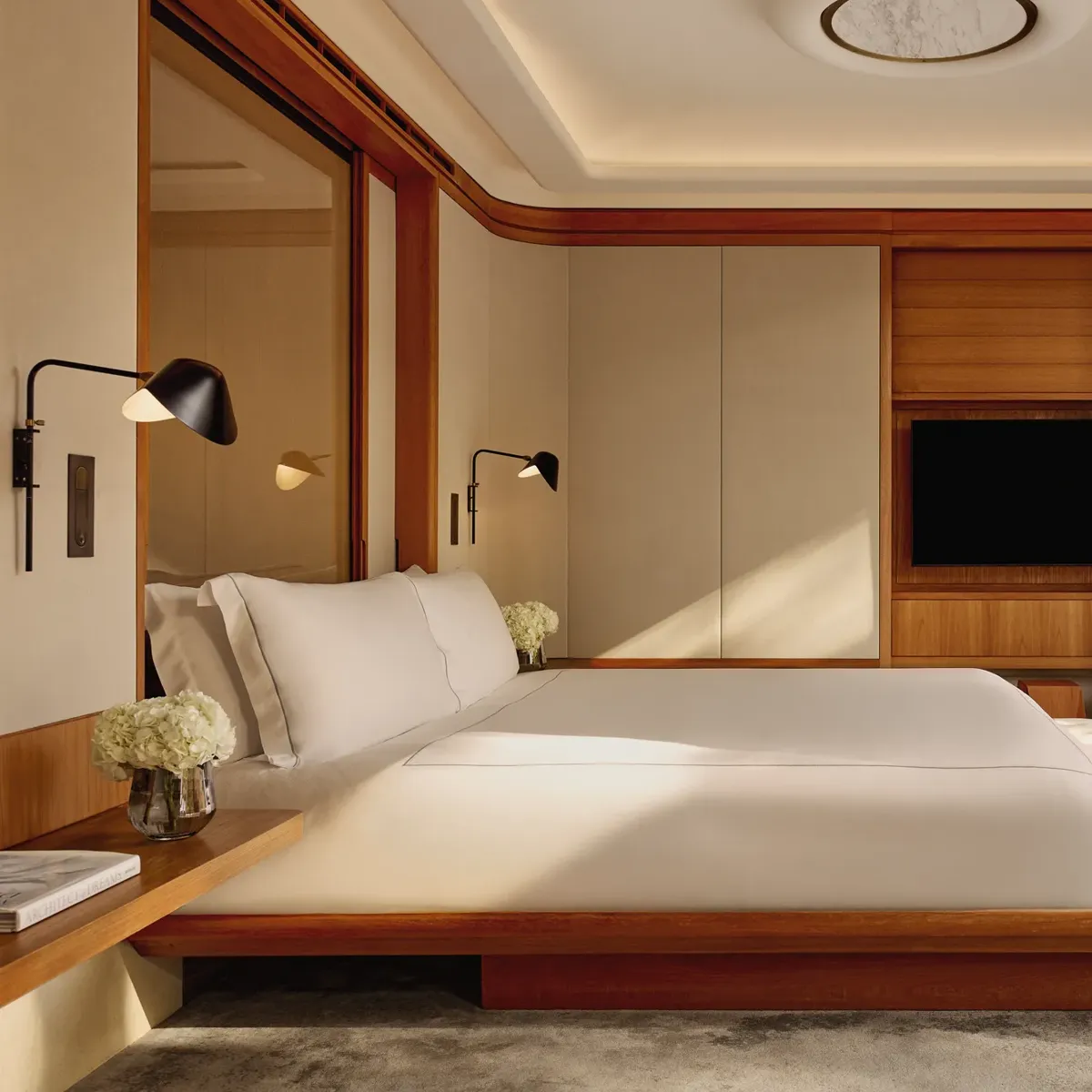 A contemporary bedroom with a low wooden bed frame and crisp white bedding, accented by wall-mounted lamps and large sliding doors opening onto a private terrace.