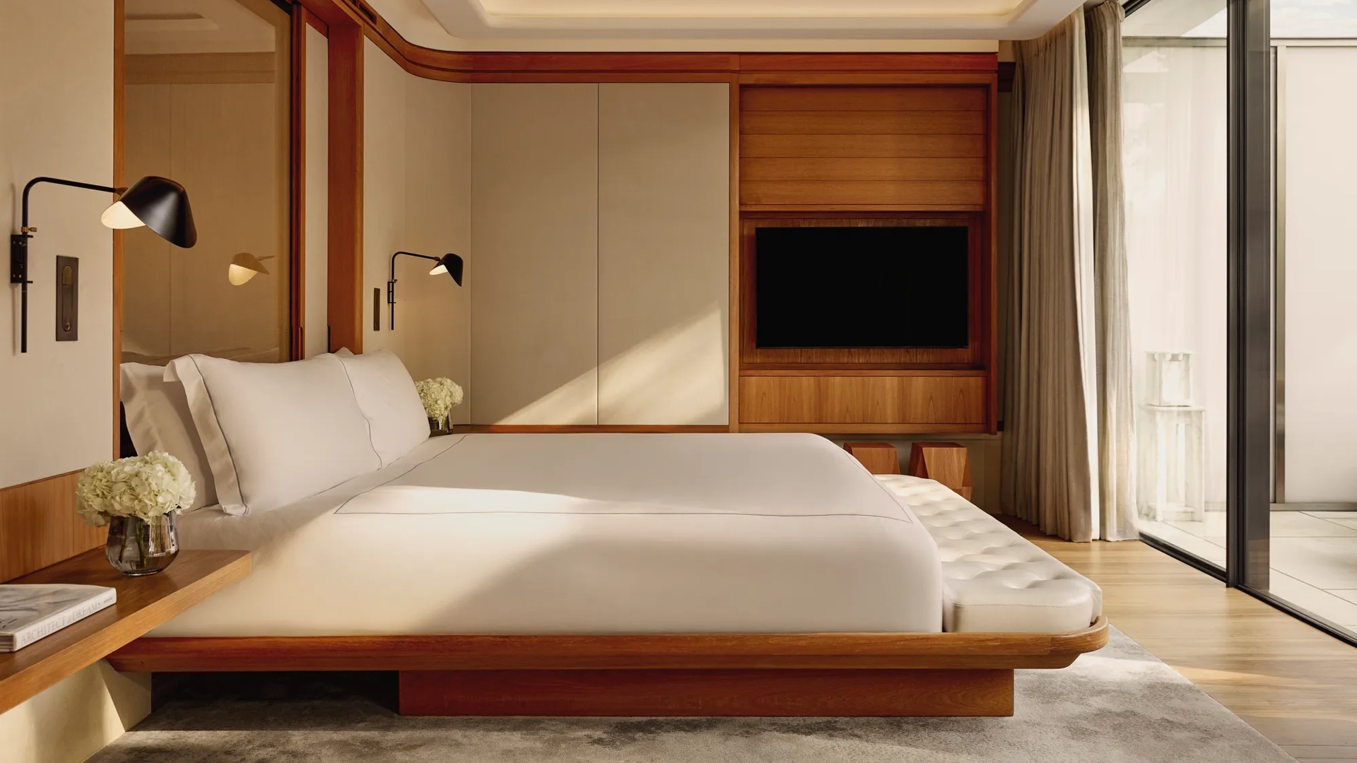 A contemporary bedroom with a low wooden bed frame and crisp white bedding, accented by wall-mounted lamps and large sliding doors opening onto a private terrace.