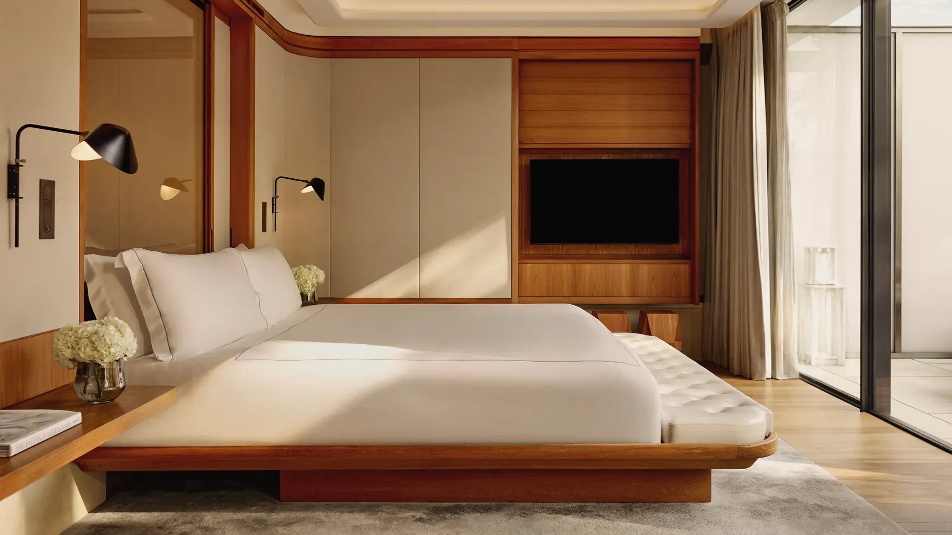 A contemporary bedroom with a low wooden bed frame and crisp white bedding, accented by wall-mounted lamps and large sliding doors opening onto a private terrace.