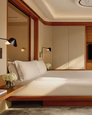 A contemporary bedroom with a low wooden bed frame and crisp white bedding, accented by wall-mounted lamps and large sliding doors opening onto a private terrace.