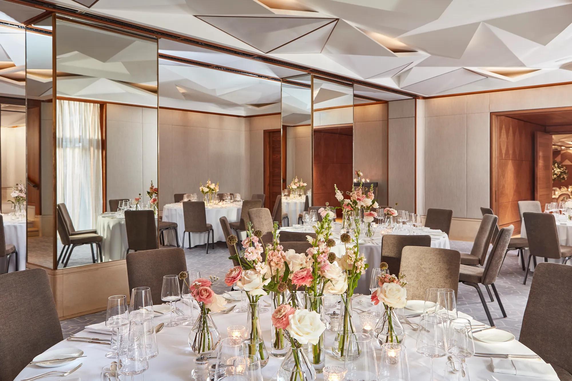 Banquet room with round tables, pink and white floral arrangements, and mirrored walls beneath geometric ceiling.