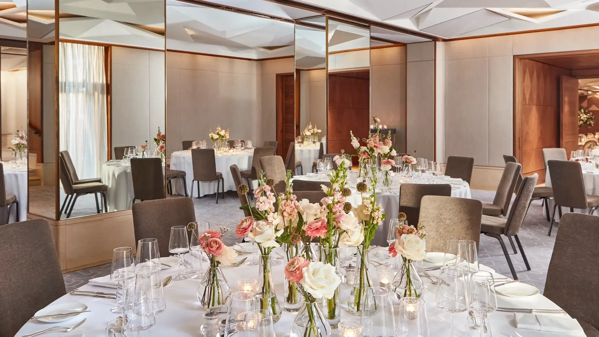 Banquet room with round tables, pink and white floral arrangements, and mirrored walls beneath geometric ceiling.