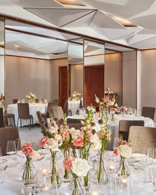 Banquet room with round tables, pink and white floral arrangements, and mirrored walls beneath geometric ceiling.