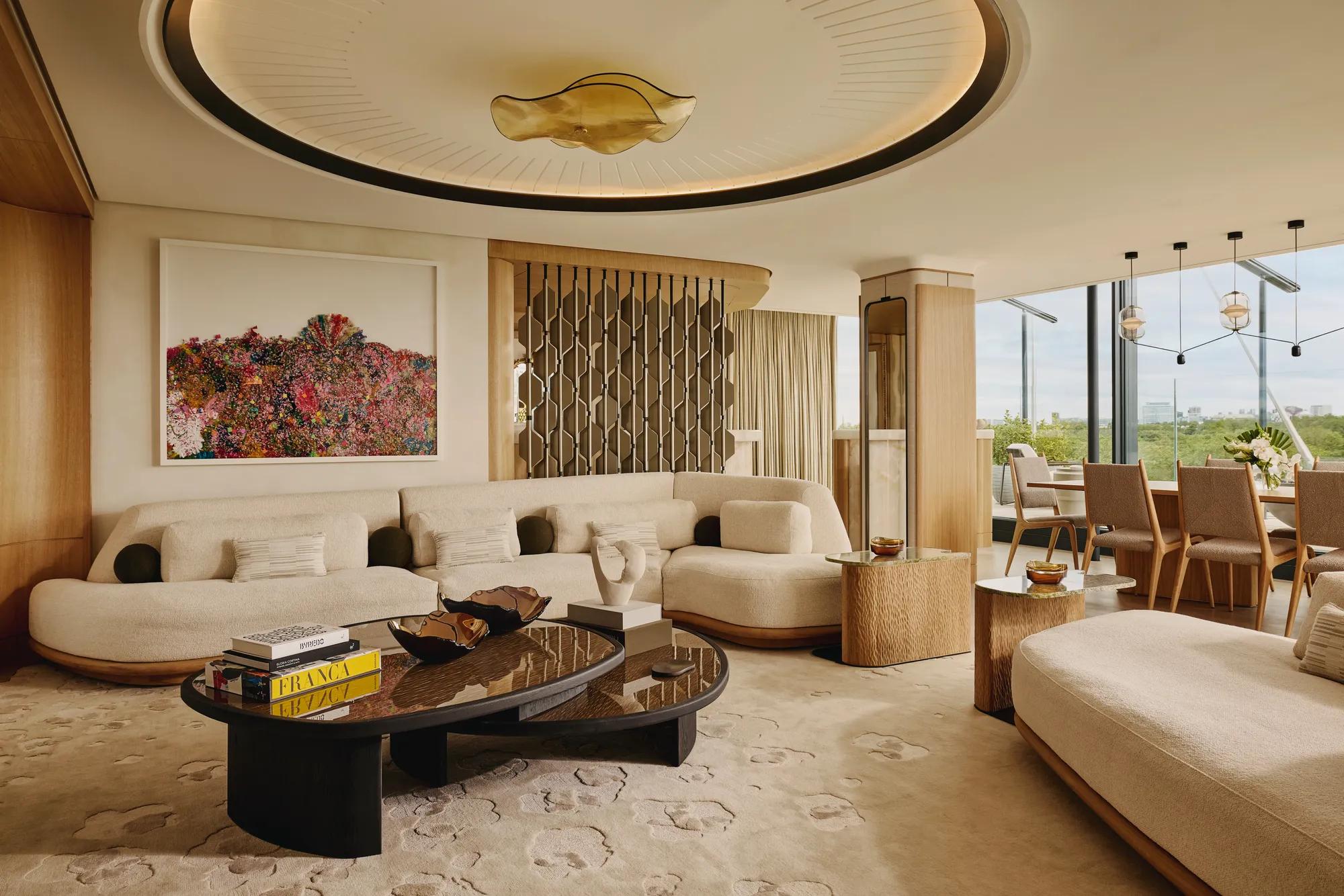 An open-plan penthouse living and dining area with cream curved sofas, sculptural wood furniture, a large contemporary chandelier, and floor-to-ceiling views of Hyde Park.