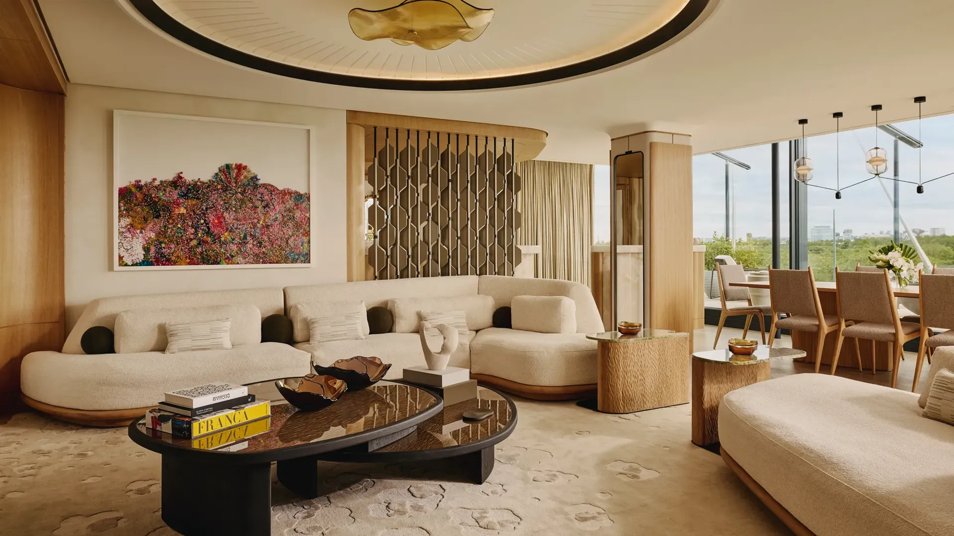 An open-plan penthouse living and dining area with cream curved sofas, sculptural wood furniture, a large contemporary chandelier, and floor-to-ceiling views of Hyde Park.