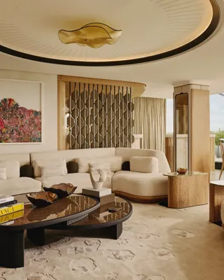 An open-plan penthouse living and dining area with cream curved sofas, sculptural wood furniture, a large contemporary chandelier, and floor-to-ceiling views of Hyde Park.