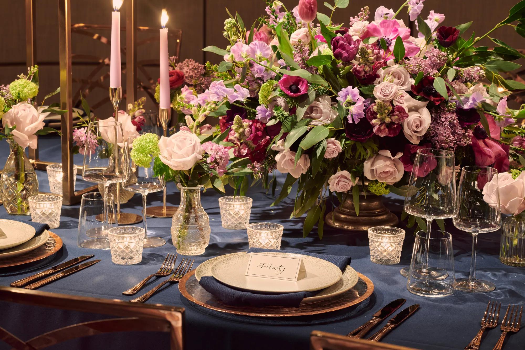Table set with blue tablecloth, pink candles, and abundant floral arrangements in pink, lilac, and burgundy tones.