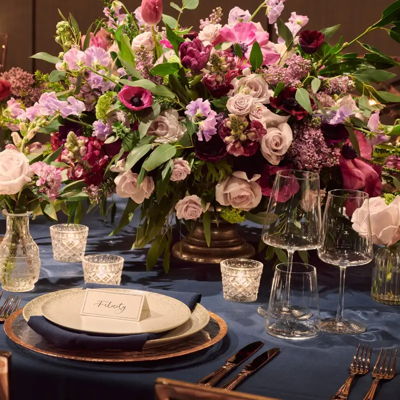 Table set with blue tablecloth, pink candles, and abundant floral arrangements in pink, lilac, and burgundy tones.