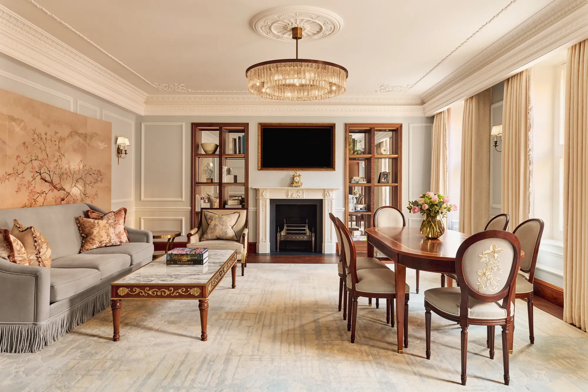 Elegant living and dining room with a grey sofa, marble-topped coffee table, and oval wooden dining table under a crystal chandelier, featuring a decorative fireplace and built-in bookshelves.