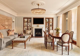 Elegant living and dining room with a grey sofa, marble-topped coffee table, and oval wooden dining table under a crystal chandelier, featuring a decorative fireplace and built-in bookshelves.
