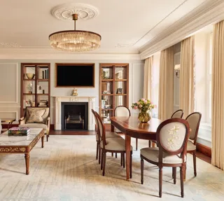 Elegant living and dining room with a grey sofa, marble-topped coffee table, and oval wooden dining table under a crystal chandelier, featuring a decorative fireplace and built-in bookshelves.