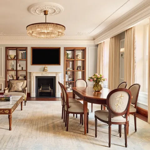 Elegant living and dining room with a grey sofa, marble-topped coffee table, and oval wooden dining table under a crystal chandelier, featuring a decorative fireplace and built-in bookshelves.