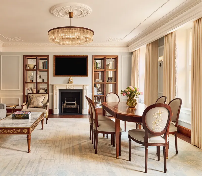 Elegant living and dining room with a grey sofa, marble-topped coffee table, and oval wooden dining table under a crystal chandelier, featuring a decorative fireplace and built-in bookshelves.