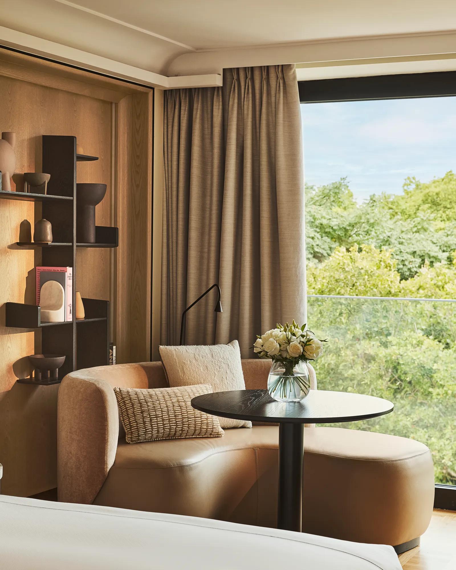 Upholstered corner seating with a round table beside a large window overlooking greenery, styled with neutral cushions and shelving.
