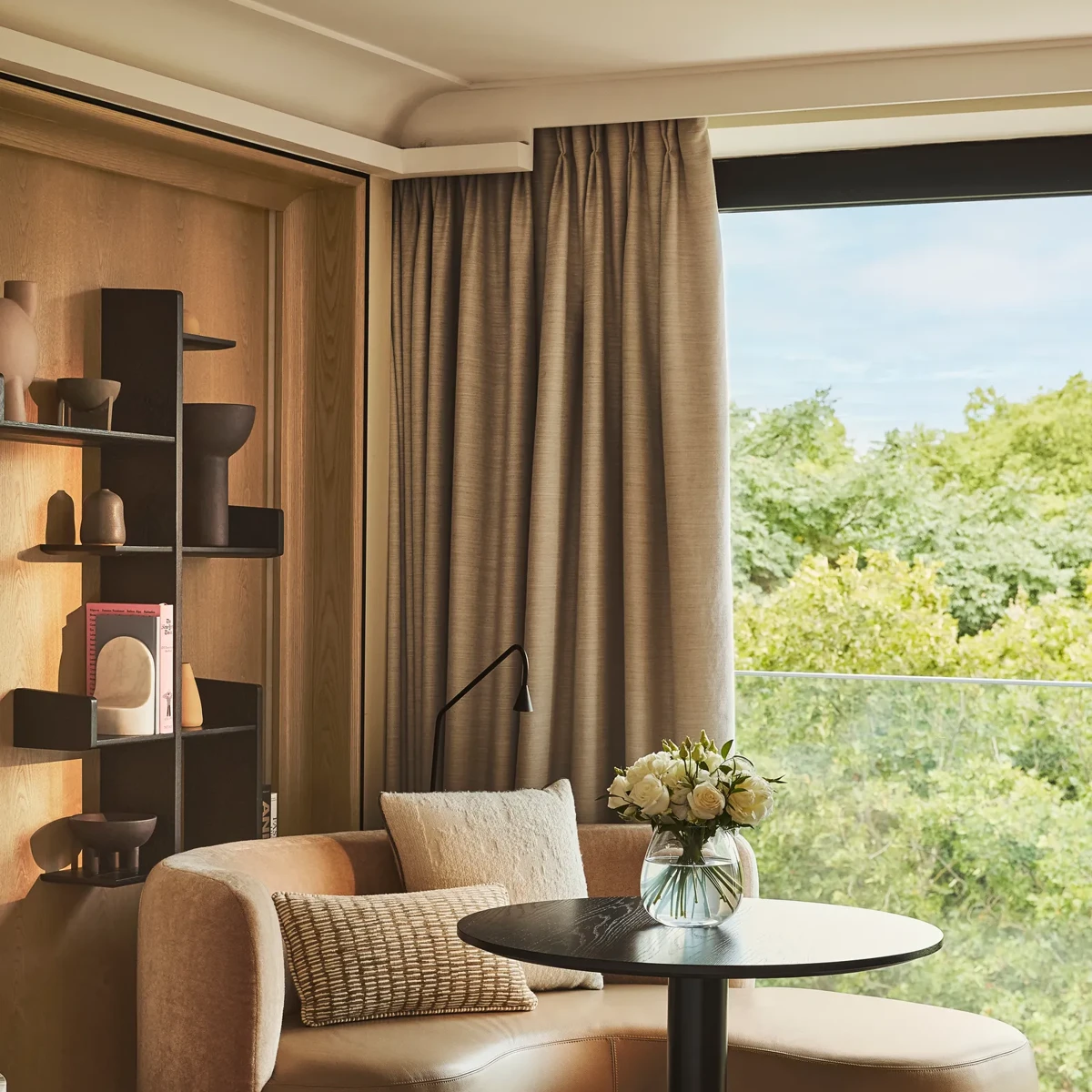 Upholstered corner seating with a round table beside a large window overlooking greenery, styled with neutral cushions and shelving.