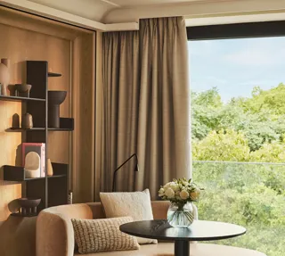 Upholstered corner seating with a round table beside a large window overlooking greenery, styled with neutral cushions and shelving.