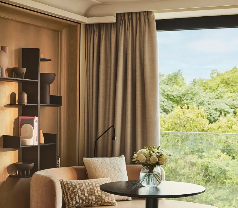 Upholstered corner seating with a round table beside a large window overlooking greenery, styled with neutral cushions and shelving.