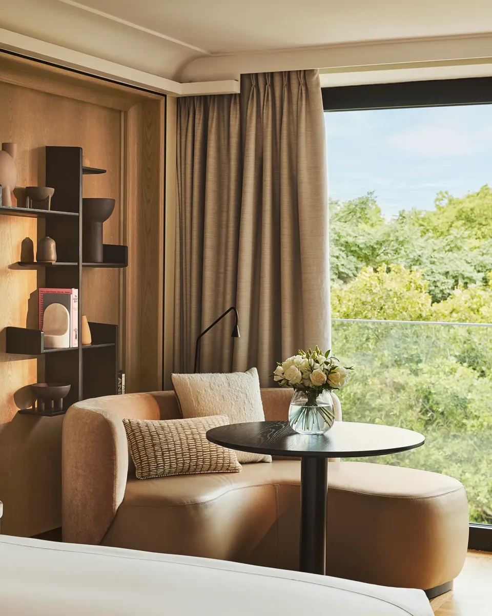 Upholstered corner seating with a round table beside a large window overlooking greenery, styled with neutral cushions and shelving.