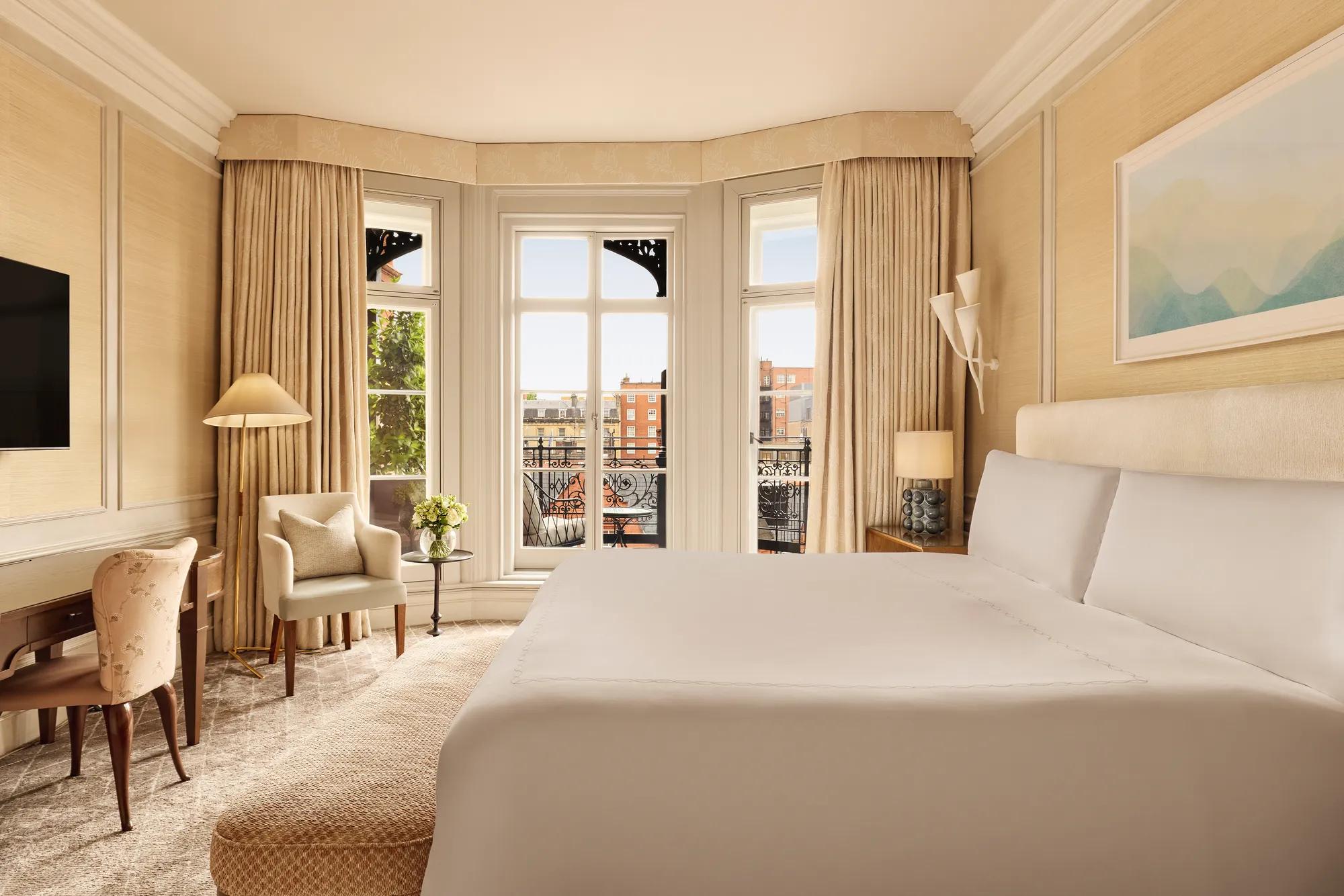 Bright bedroom with a large bed, writing desk, and armchair by tall bay windows draped in golden curtains, opening to a balcony with views of red-brick buildings.